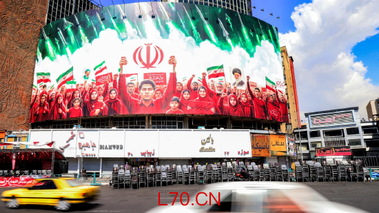 2026 年 4 月 6 日，德黑兰瓦利亚斯广场，车主驾车经过一幅大型政治宣传横幅，画面展示伊朗示威者身后发射导弹，以示对政府的支持。当日，以色列空袭击毙伊朗革命卫队情报主管；伊朗则无视美国总统威胁 —— 若不重新开放霍尔木兹海峡，将摧毁伊朗民用基础设施。